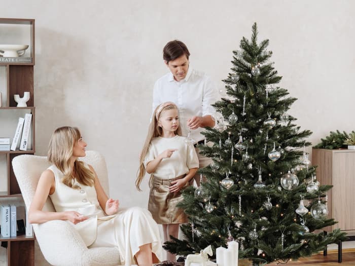Mom sits in a bright armchair with a mug while dad and daughter decorate the artificial Christmas tree FairyTrees FT14 with silver ornaments in the living room.