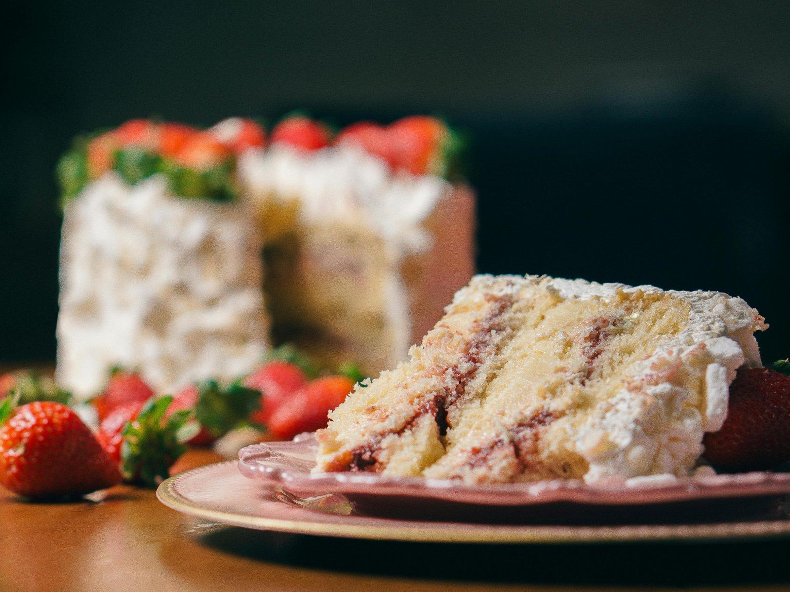 A slice of Christmas cake with cream and strawberries is placed on a plate, with the whole decorated cake visible in the background.