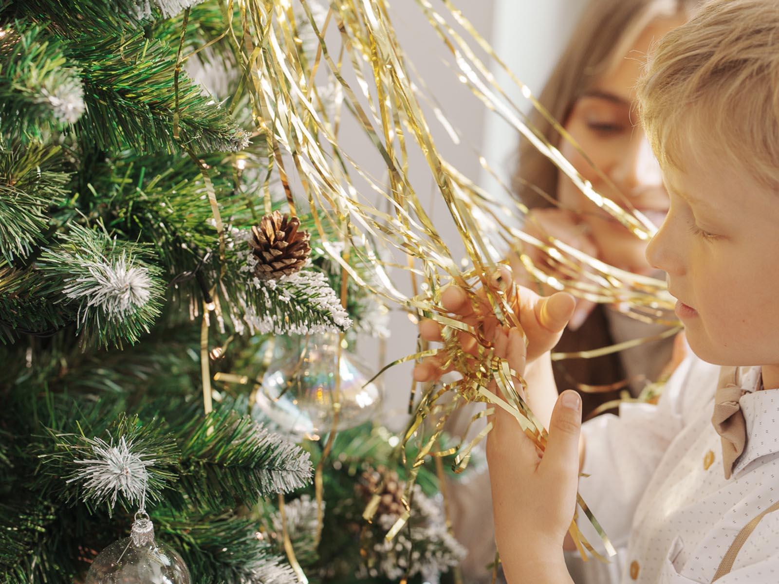 Son places golden tinsel on the artificial Christmas tree FairyTrees FT04, mom helps in the background, and the close-up shows snowy branches, pinecones, and glass baubles.