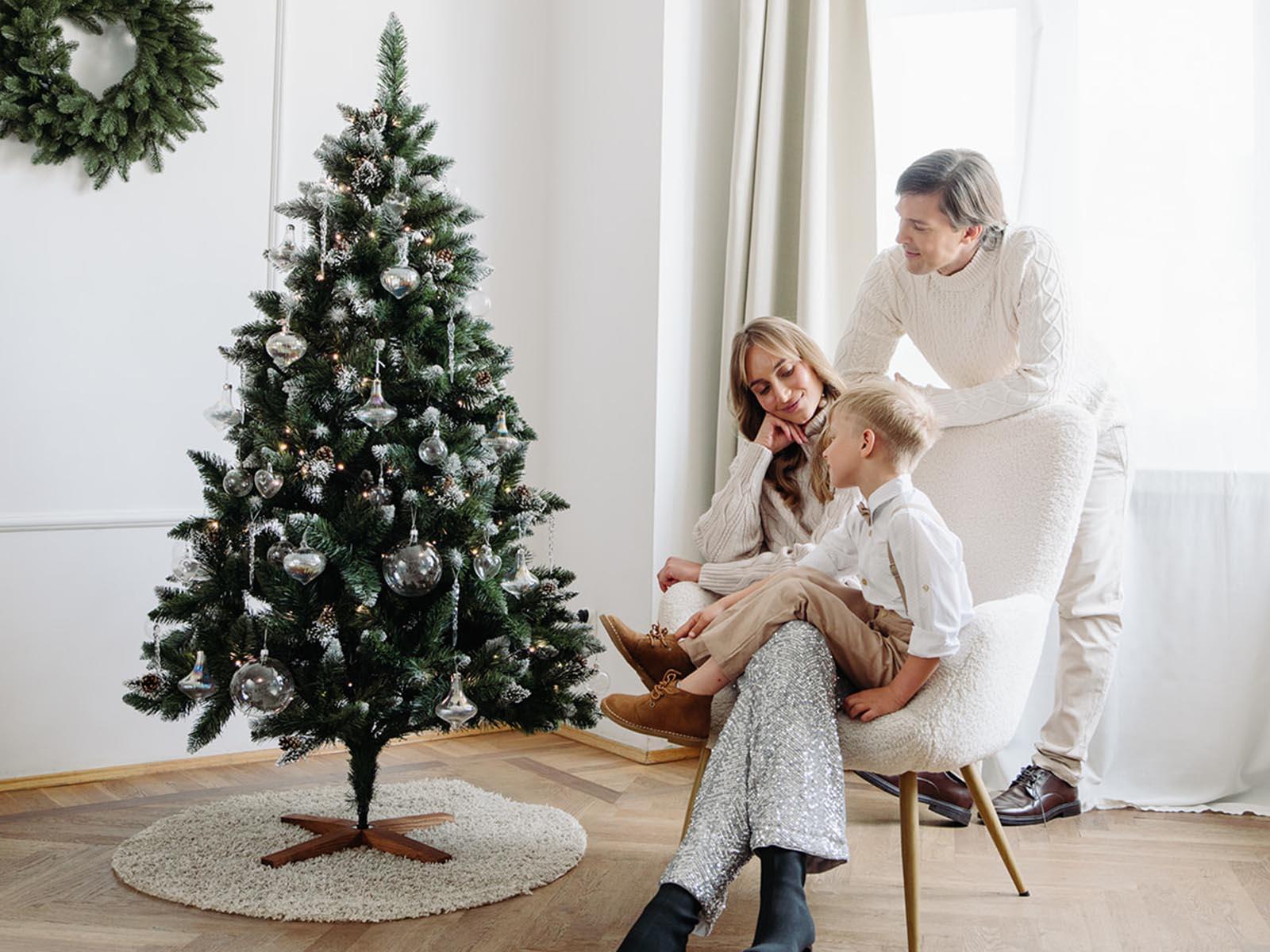 Mom relaxes in an armchair with her son, dad leans toward them, and a snow-dusted artificial Christmas tree FairyTrees FT04 glows with warm lights and glass ornaments.