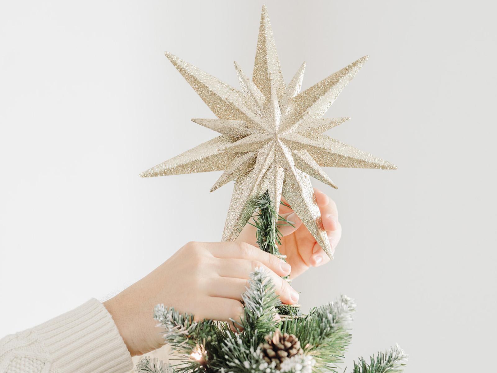 Hands place a glittering star on top of the artificial Christmas tree FairyTrees FT04 with snowy needles, lights, and pinecones.