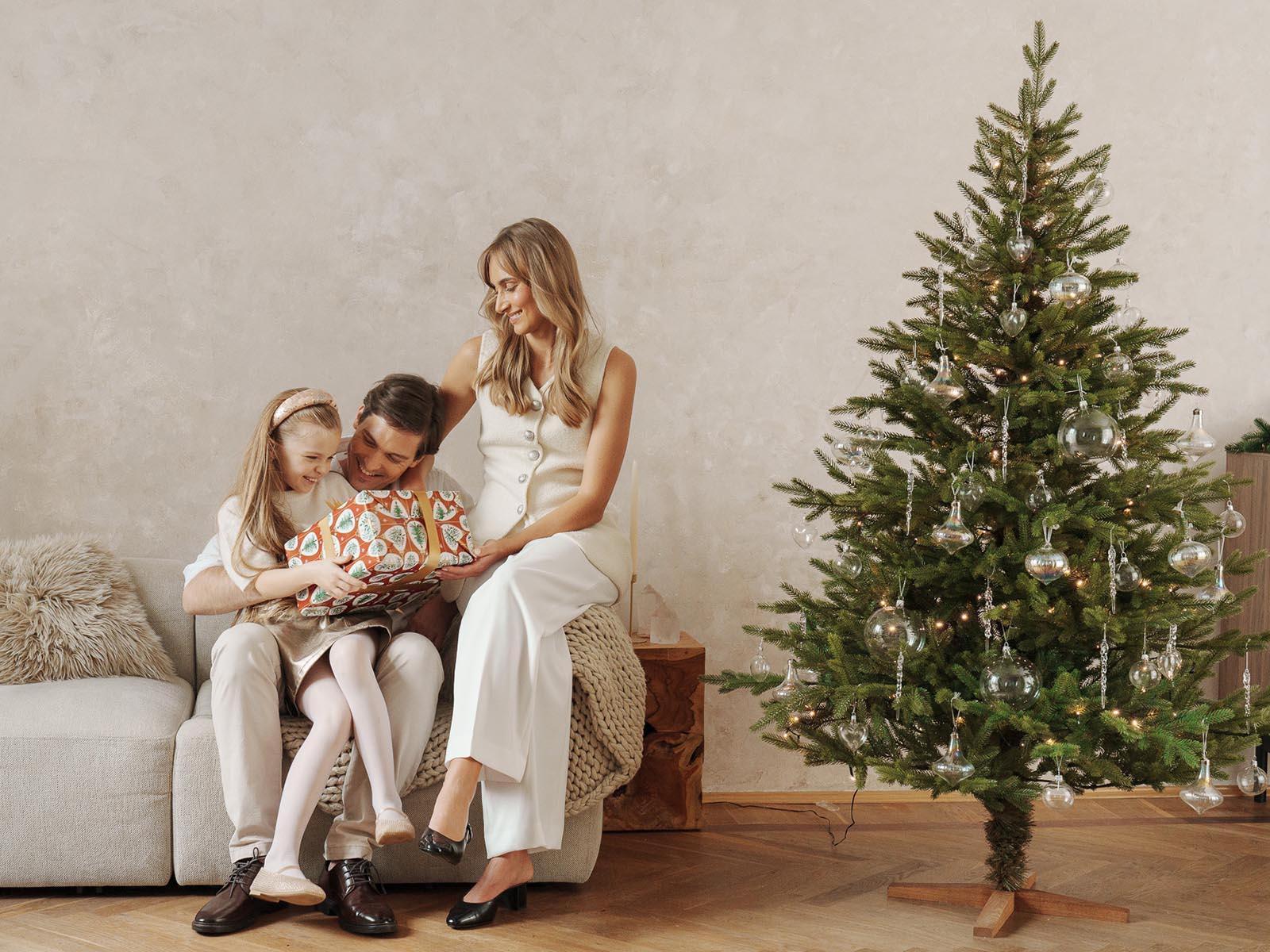 Mom and dad sit with their daughter on the sofa opening a gift, while the artificial Christmas tree FairyTrees FT06 shines with silver decorations nearby.