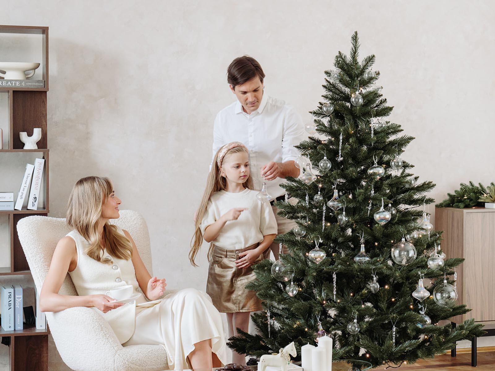 Mom sits in a bright armchair with a mug while dad and daughter decorate the artificial Christmas tree FairyTrees FT14 with silver ornaments in the living room.