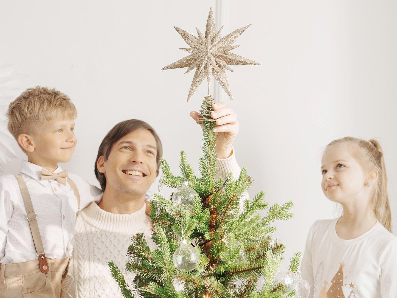 Dad places a star on top of the FairyTrees FT32 artificial Christmas tree, accompanied by his children.