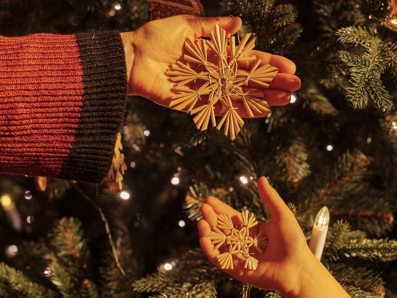The hands of a grandmother and her granddaughter hold straw stars next to a lit FairyTrees FT29 artificial Christmas tree.