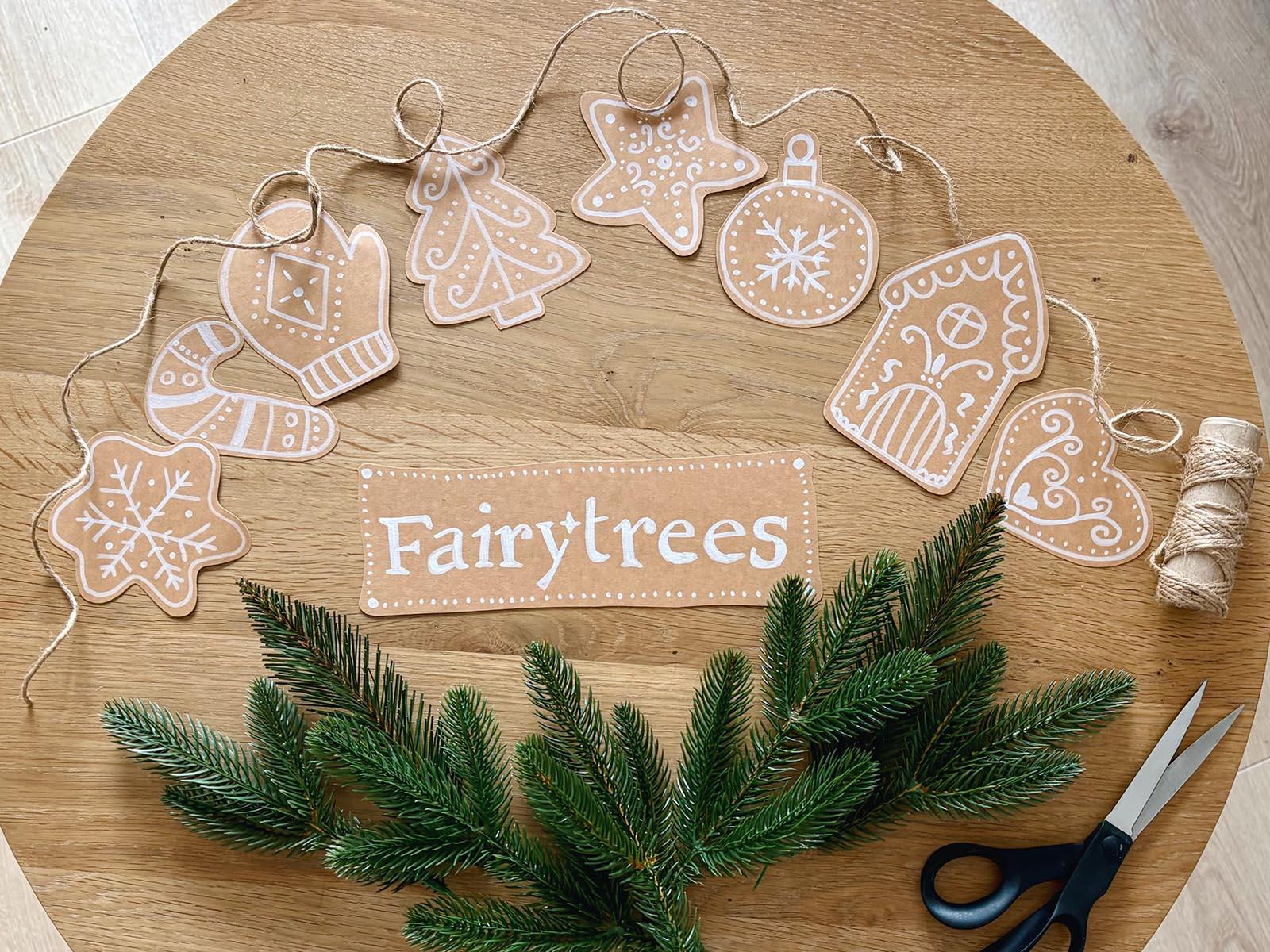 The branches of the FairyTrees artificial Christmas tree lie on a wooden tabletop alongside gingerbread-style paper decorations, ready to be turned into garlands.