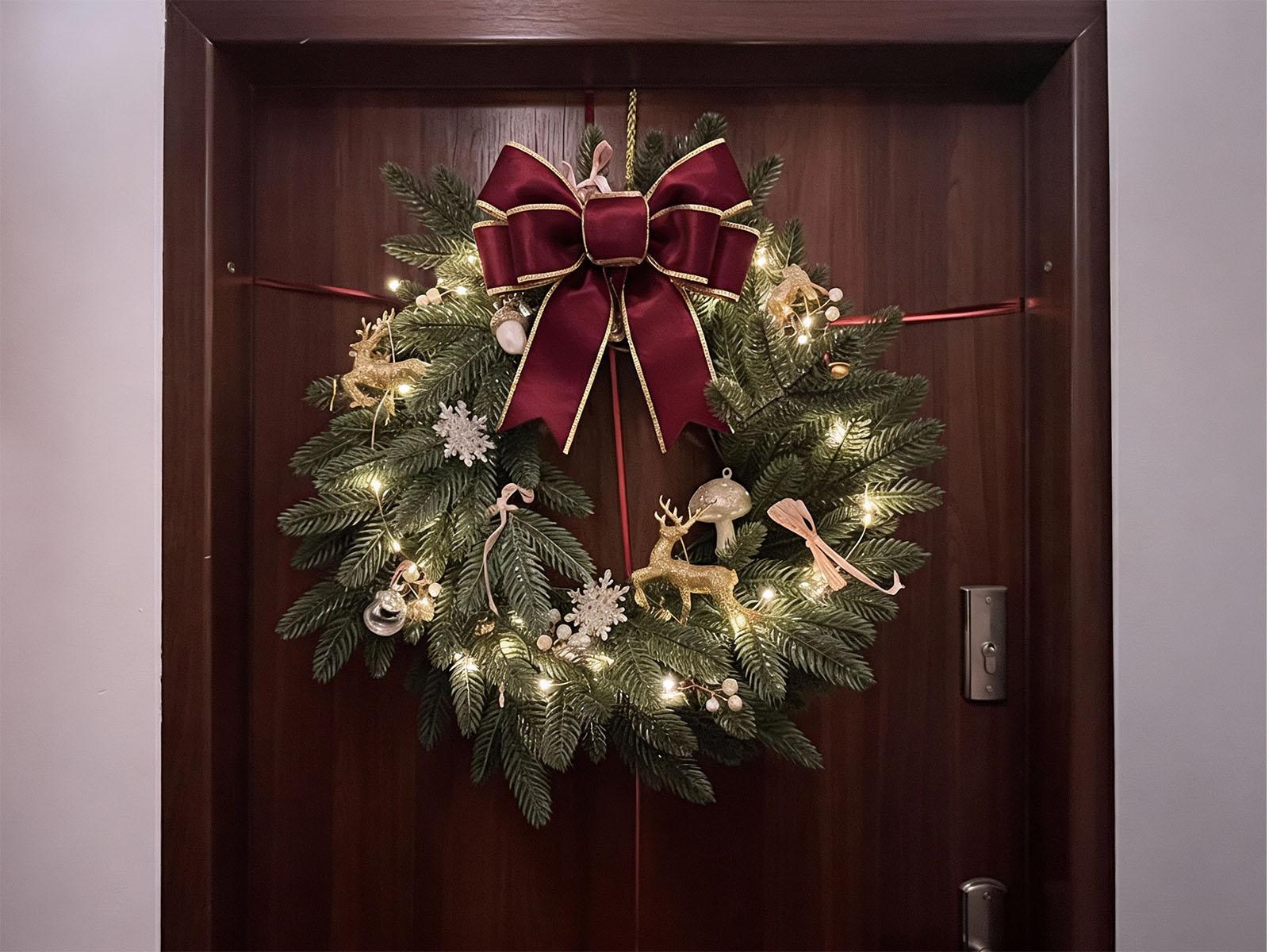 A Christmas wreath made from branches of an artificial Christmas tree hangs on a dark door, adorned with lights, a golden reindeer and a large burgundy bow.
