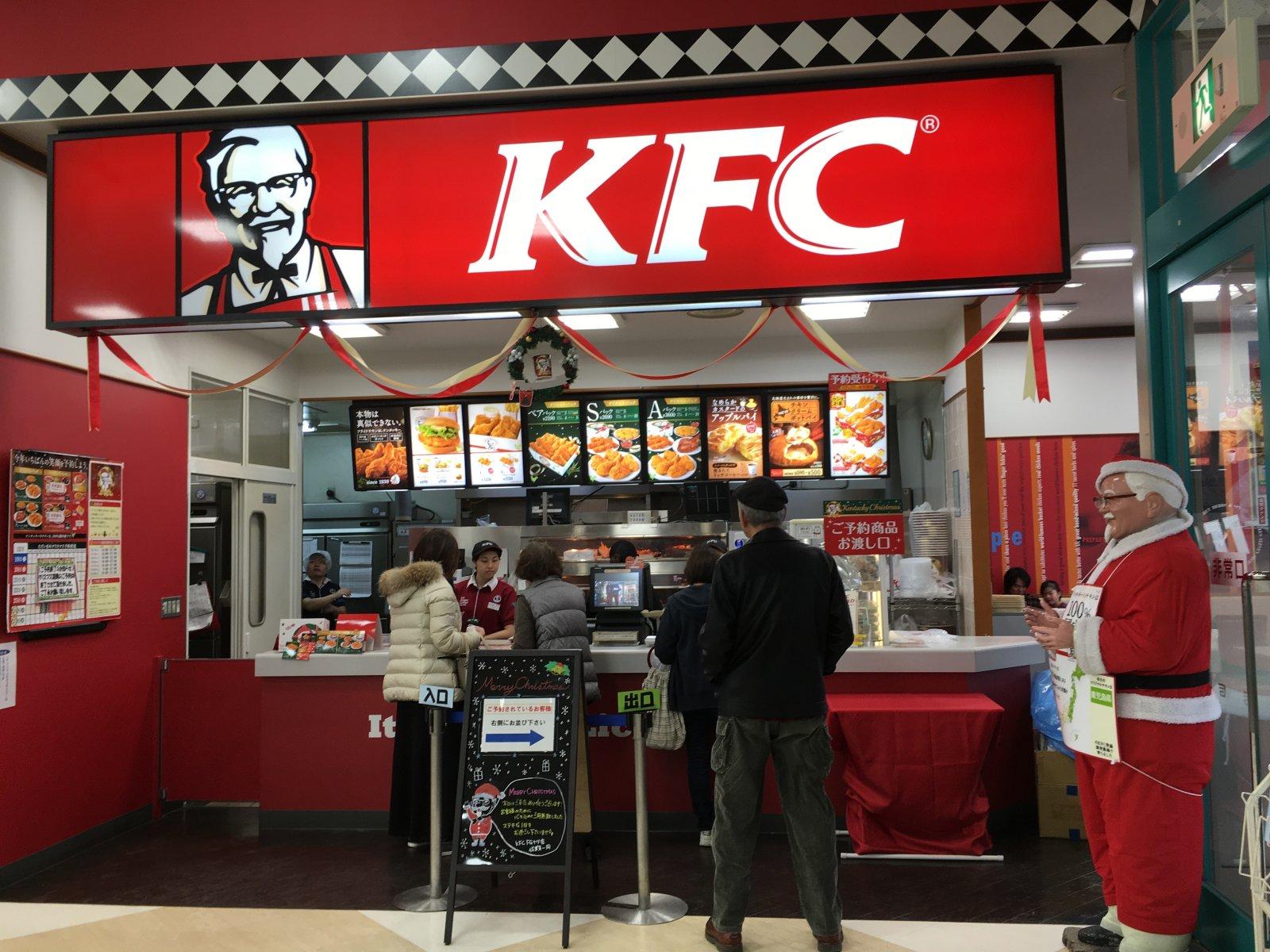 Customers order food at the counter of a Christmas-decorated KFC restaurant, with a person dressed as Santa Claus standing nearby.