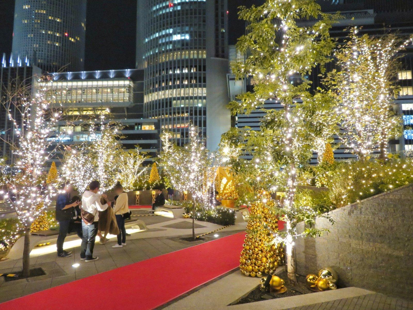 A rooftop terrace of the Dai-Nagoya building in Tokyo is decorated with festive lights and Christmas trees, surrounded by modern city buildings.